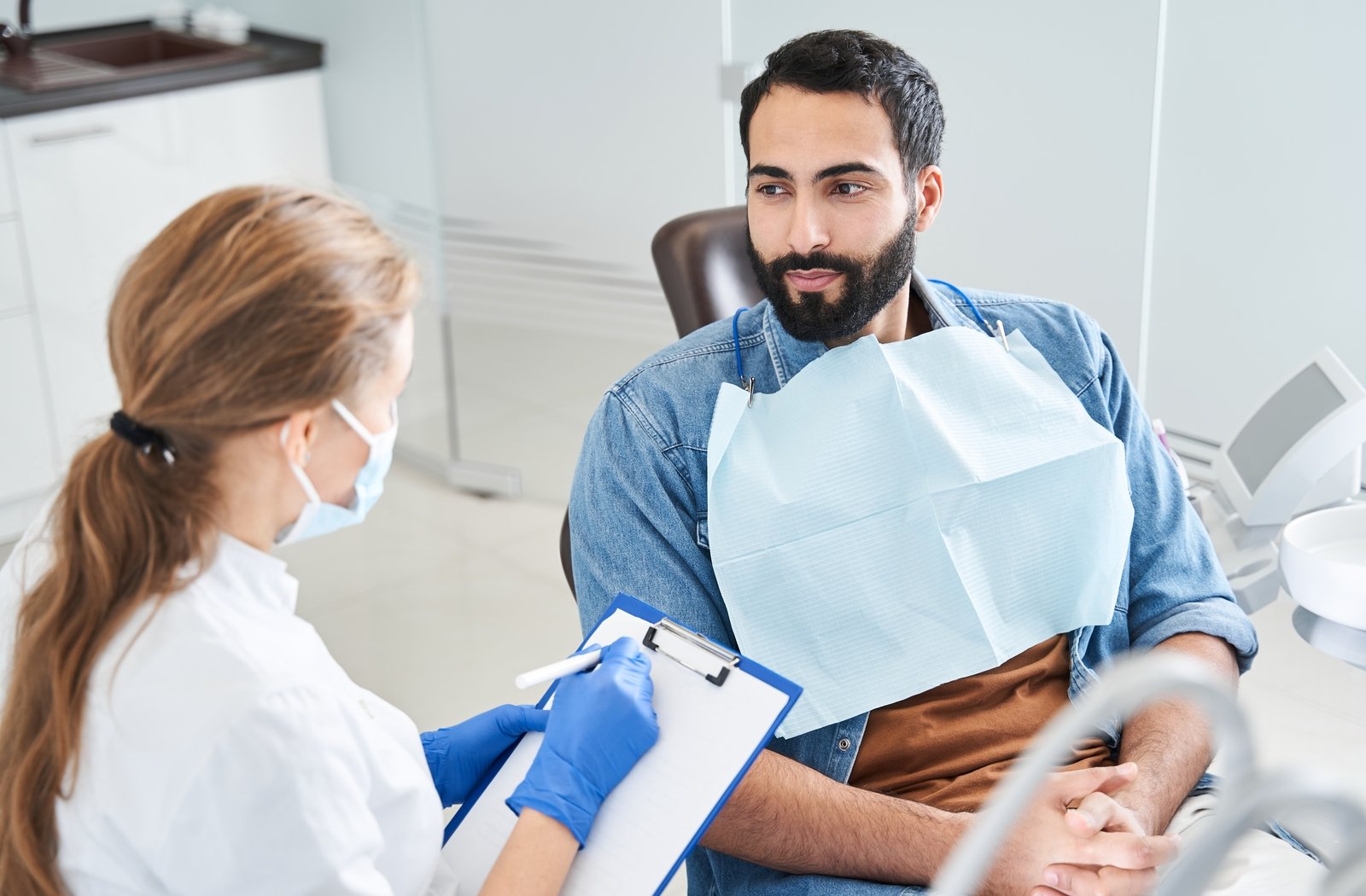 A man in a dentist's chair having a conversation with his dentist that is holding a clip board writing something down