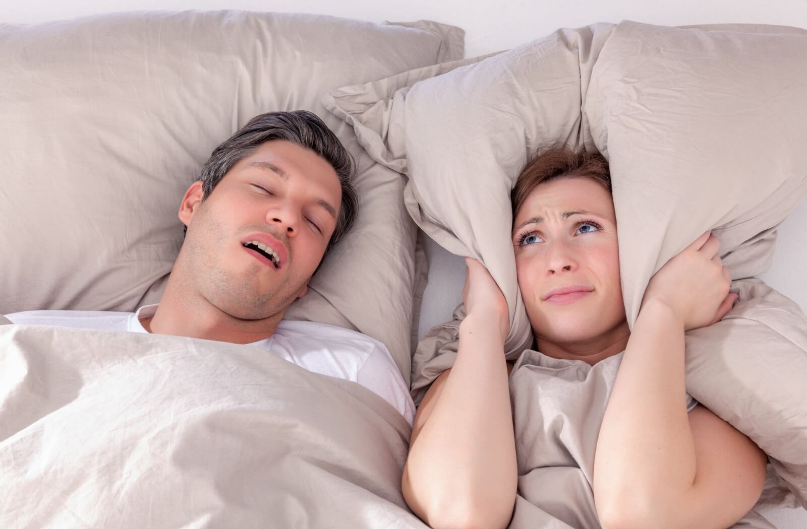 A husband and wife sleeping, the wife is covering her ears due to a loud snoring sound of her husband with apnea.