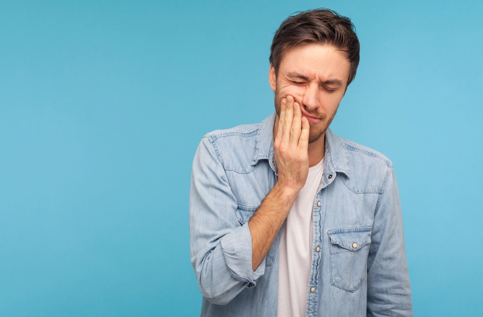 A man standing against a blue background and holding his right hand to his cheek with a pained expression