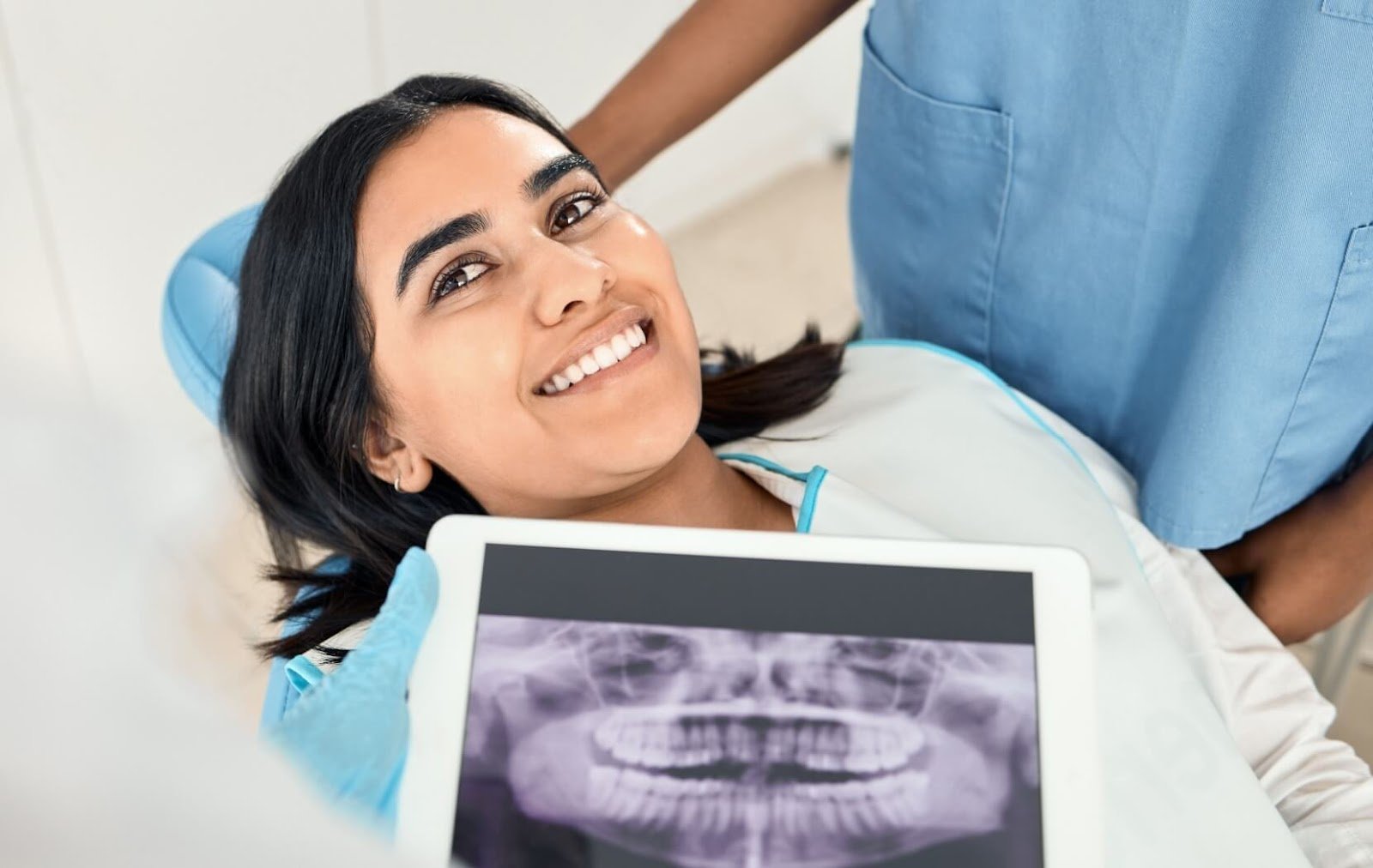 A dental patient smiles as their dentist holds up an x-ray taken after they are able to undergo a successful dental procedure.